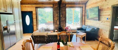 A wooden cabin interior with a dining table, chairs, a stone pillar, a ceiling fan, and a kitchen area.