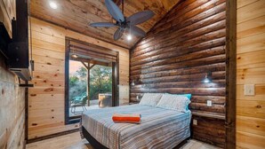 A log cabin bedroom with a bed, a ceiling fan, and a view of the outdoors. - Hidden Hills Hideaway- Broken Bow/Hochatown- 2bd (Smithville)