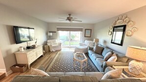 A spacious living room with a large sofa, a coffee table, and a television mounted on the wall. There is a ceiling fan and a window providing natural light. - Oceanview 2 bedroom Flat unit 137 (St. Augustine)