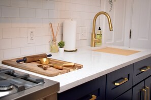 A modern kitchen with white tiles, a stainless steel stove, a wooden cutting board, and a gold faucet.