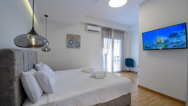 A modern bedroom with a large bed, a hanging light fixture, a flat-screen TV, and a view of the outdoors through a window. - Lush Sapphire apt. steps from the Acropolis! (Athina)