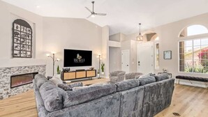 Moderni olohuone, jossa on suuri kulmasohva, takka, litteänäyttöinen televisio ja kattotuuletin.