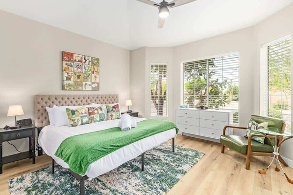 A bedroom with a bed, a green blanket, a headboard, a nightstand, a lamp, a window with blinds, a chair, and a ceiling fan. - Tatum Ranch Retreat | Heated Pool & Backyard Fun (Cave Creek)