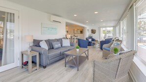 Un salón amplio con un sofá gris, sillones azules y una silla de mimbre. Hay una mesita con una lámpara y una planta. La habitación tiene ventanas grandes y vistas al exterior.