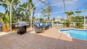 Un patio al aire libre con piscina, parrilla y zonas de estar, rodeado de plantas tropicales.
