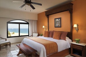 A hotel room with a large bed, a ceiling fan, a view of the beach, and a painting on the wall.