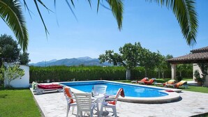 A swimming pool with lounge chairs and a table, surrounded by a stone patio and greenery, with mountains in the background. - Finca Burga, vacation home in a quiet location, views, pool, A/C, Wifi (Puerto Pollenca)