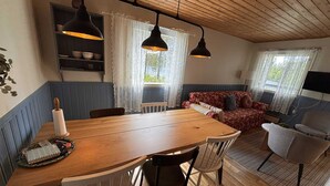 Une pièce chaleureuse avec une table à manger en bois, des chaises, un canapé rouge à motifs et un fauteuil gris. Deux lampes suspendues sont placées au-dessus de la table, et une étagère contient des bols et un vase.