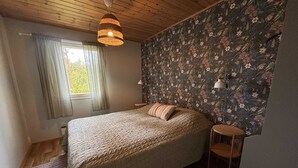 Une chambre à coucher avec un lit, une fenêtre avec des rideaux, un mur recouvert d’un papier peint à motifs floraux, un luminaire suspendu et une petite table de chevet.