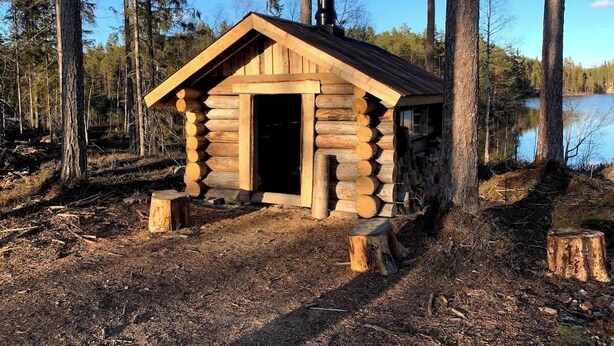 Une cabane en rondins avec une cheminée, entourée d’arbres et bordée par un plan d’eau.