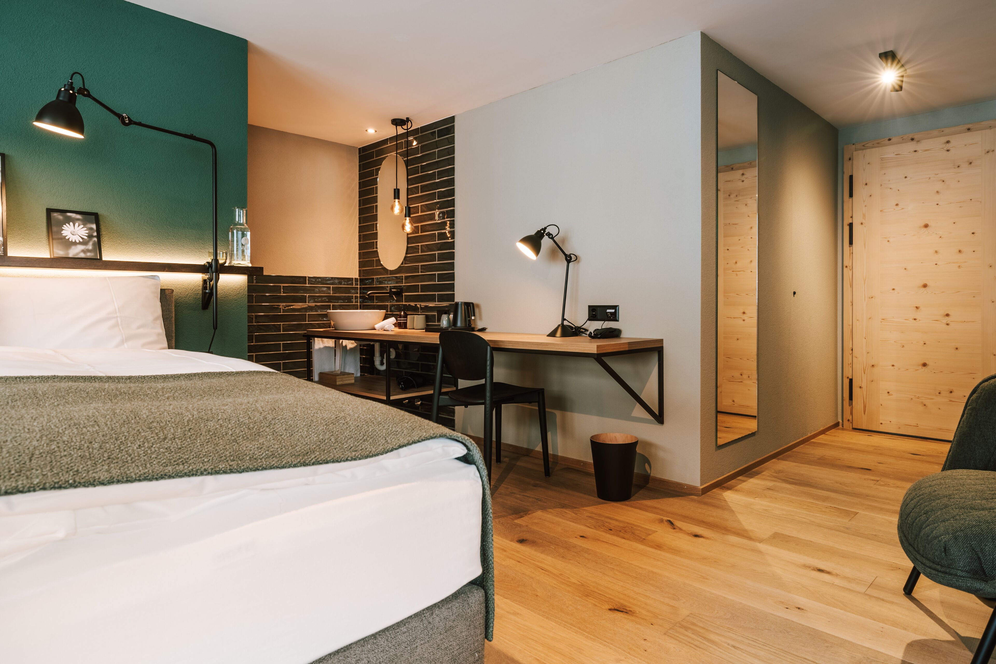 A modern hotel room with a bed, desk, and chair, a small kitchen area, and a wooden door.