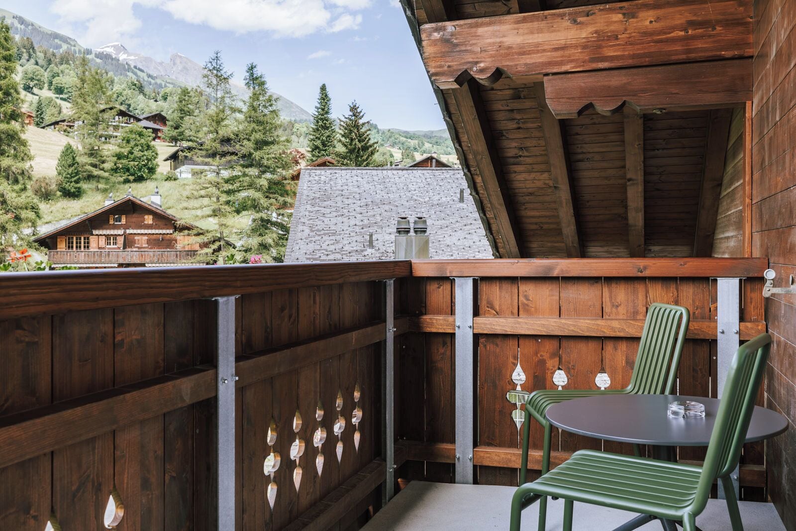 A wooden balcony with a table and chairs, overlooking a mountainous landscape with several wooden buildings.
