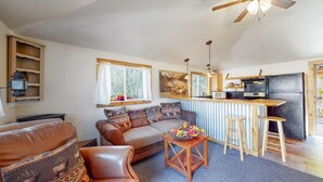 A cozy living room with a sofa, a coffee table, and a kitchen area with a refrigerator and a bar. - Take it EZ cabin overlooking the river with HOT TUB  on the deck 5 min to Estes! (Drake)