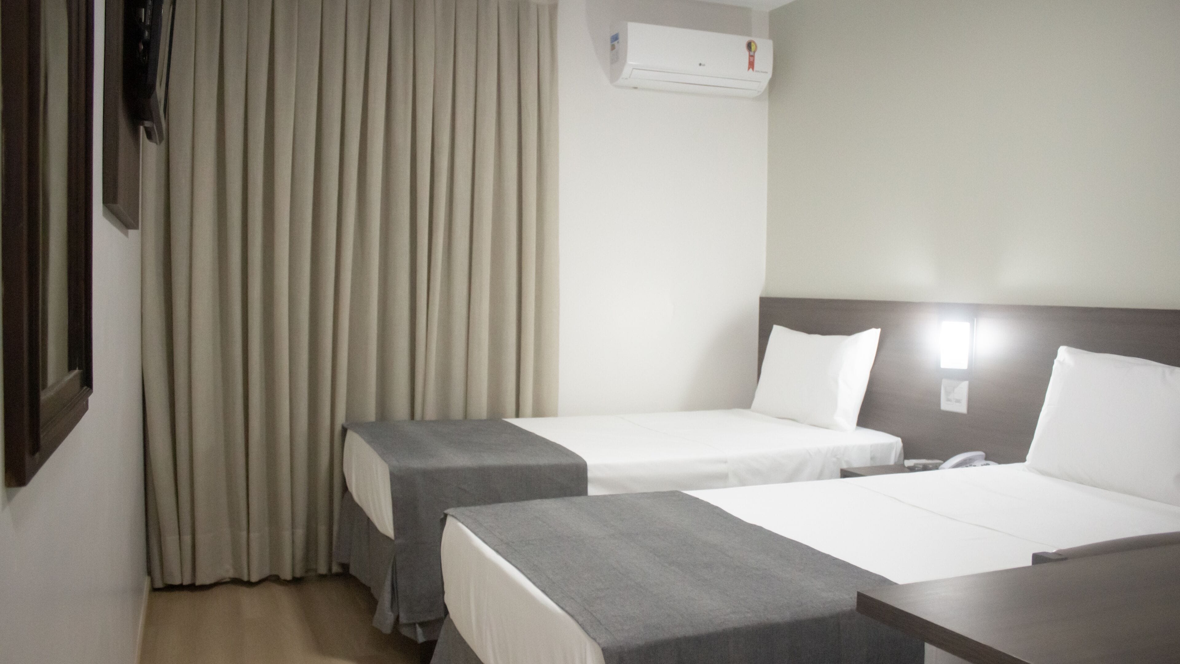Habitación de hotel con dos camas, una ventana con cortinas, un equipo de aire acondicionado y una mesita de noche de madera.