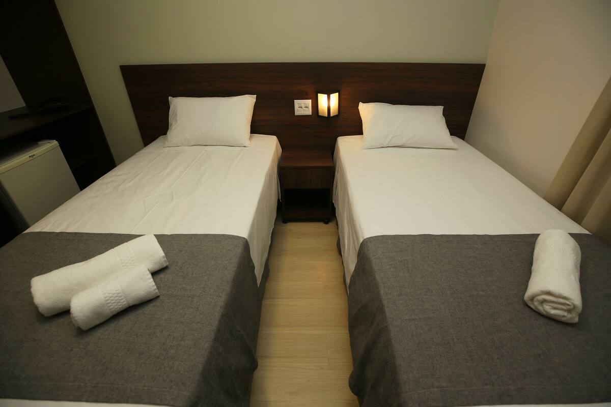 Ein Hotelzimmer mit zwei Einzelbetten, jeweils mit einem Kopfkissen und einem zusammengerolltem Handtuch. Zwischen den Betten befindet sich ein Nachttisch mit einer Lampe.