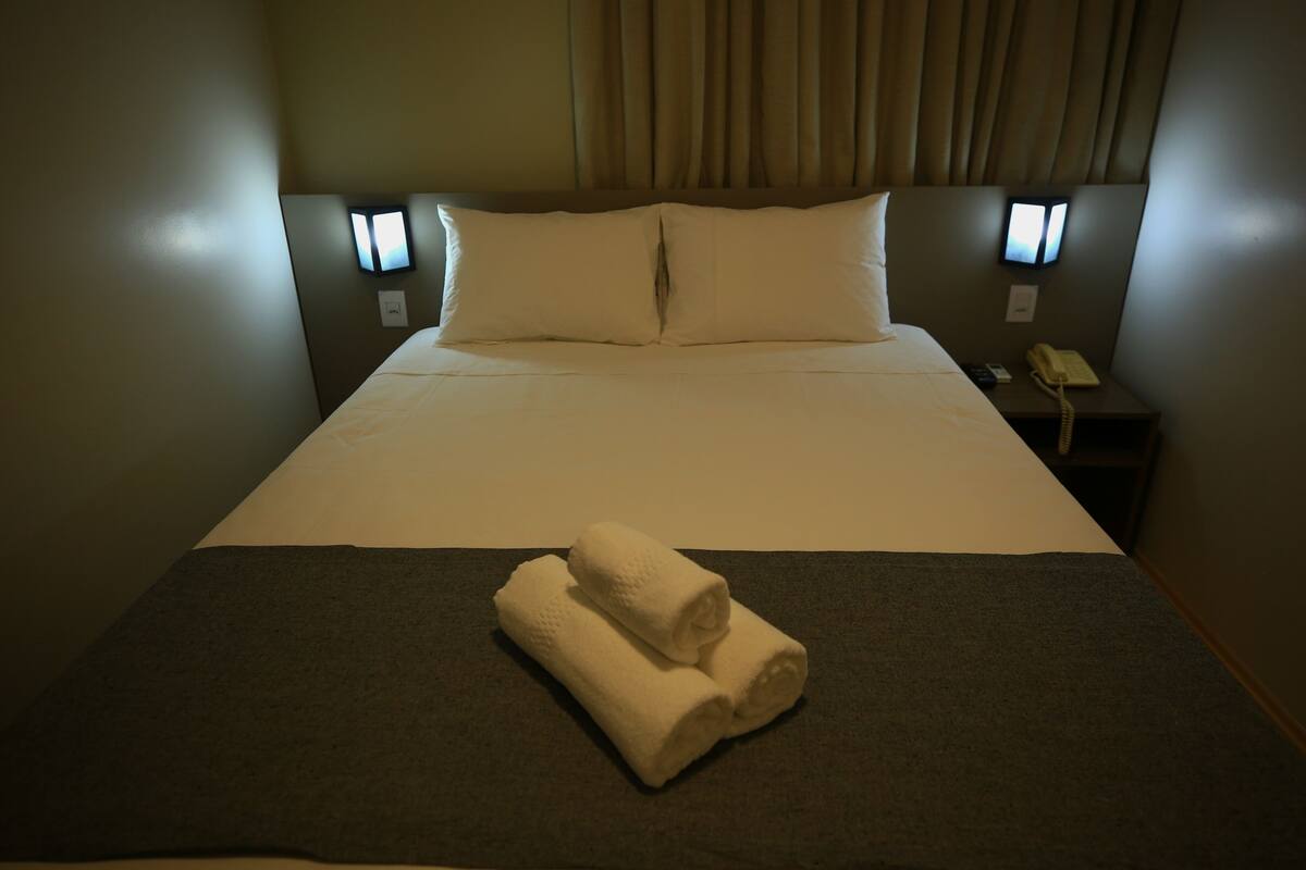 Ein Hotelzimmer mit einem ordentlich bezogenen Bett, zwei Kissen, einem Nachttisch mit Telefon und einer Wandleuchte.