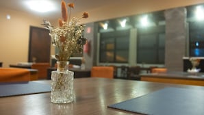 A dining table with a clear glass vase containing dried flowers. The room has a modern design with wooden furniture and large windows.