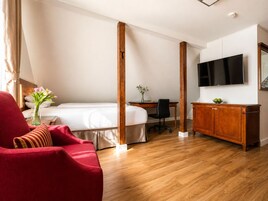 Et hotelværelse med en seng, en rød lænestol, et træskrivebord, et fjernsyn og et træskab.