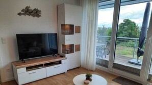 A modern living room with a flat-screen TV on a wooden console, a white sofa, two round tables, and a balcony with outdoor furniture. - Modern top floor apartment with a view of the sea and Stralsund (Altefähr)