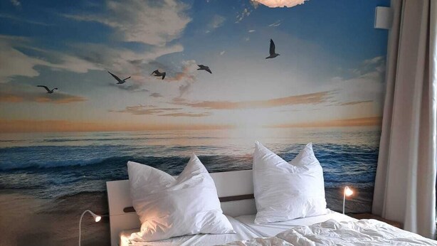 A bedroom with a mural of a sunset over the ocean, a bed with white bedding, a bedside lamp, and a hanging light fixture. - Modern top floor apartment with a view of the sea and Stralsund (Altefähr)