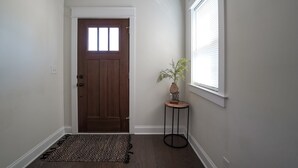 Un pasillo con una puerta de madera, una pequeña mesa redonda con una planta y una ventana con persianas.
