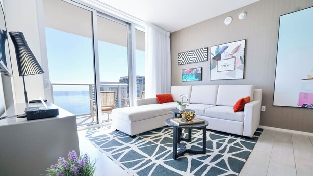 Ein modernes Wohnzimmer mit einem weißen Sofa, einem kleinen runden Tisch und einem gemusterten Teppich. Ein großes Fenster bietet Blick auf Gebäude und einen Balkon.