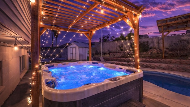 A hot tub under a wooden pergola with string lights, surrounded by a backyard with a pool.