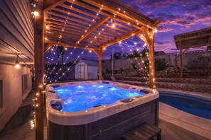 Uma banheira de hidromassagem sob uma pérgola de madeira com luzes de cordão, rodeada por um quintal com piscina.
