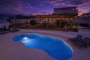 A swimming pool with a pergola and string lights, surrounded by lounge chairs and a stone wall.