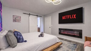 A modern bedroom with a bed, a fireplace, a mounted TV displaying "NETFLIX", and a door leading to a bathroom.