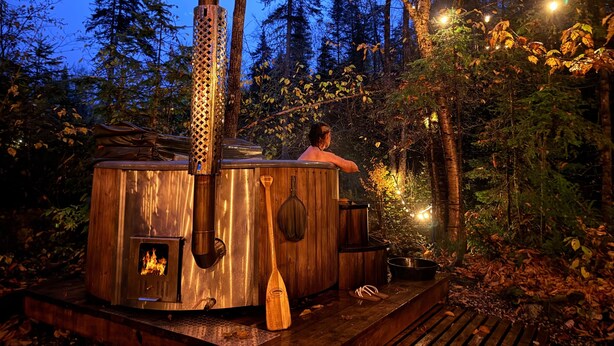Una sauna de leña en un entorno boscoso con una chimenea grande, un banco de madera y una pequeña mesa con una olla.
