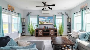 A living room with a ceiling fan, a wooden entertainment center, a sofa, a coffee table, and large windows with curtains. - Steps to Surfside Beach | Games, Views & Pets (Surfside Beach)