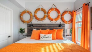 A bed with an orange quilt, a 'Welcome' pillow, and four life rings on the wall. - Steps to Surfside Beach | Games, Views & Pets (Surfside Beach)