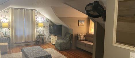 A cozy attic bedroom with a bed, a chair, a TV, and a fan.