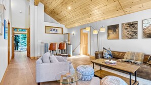 A spacious living room with a high ceiling, wooden beams, a sofa set, a coffee table, and a dining table. There is a staircase leading to another level, and a door leading to a balcony with a view of trees. - Lake Views, Hot Tub, Walk to Beach! (Carnelian Bay)