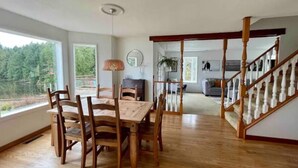 Un comedor amplio con una mesa y sillas de madera, un ventilador de techo y vistas a un área boscosa a través de grandes ventanales.