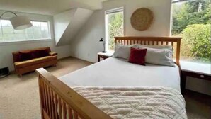 Un dormitorio con techo inclinado, una cama con cabecera de madera, una mesita de noche, una lámpara, una ventana con vistas a zonas verdes y un pequeño sofá.