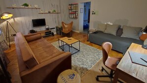 Ein modernes Wohnzimmer mit einer Couch, einem Couchtisch und einem Fernseher. Es gibt Regale mit Dekoration und einen Sessel.