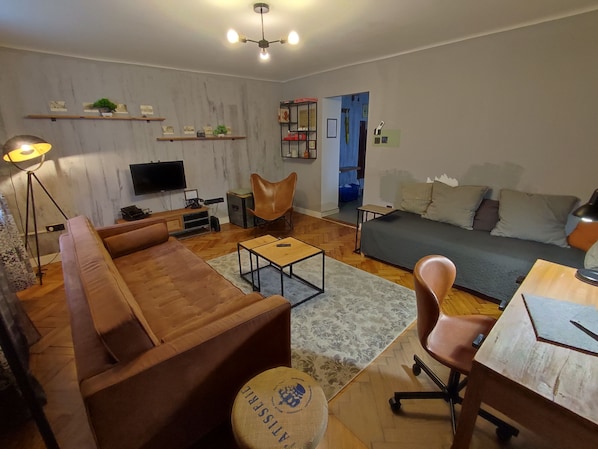 A modern living room with a sofa, coffee table, and a TV. There are shelves with decorative items and a chair.