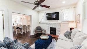 A living room with a fireplace, a television mounted above it, and a ceiling fan. There is a dining area visible through an open doorway. - Our Nest at Spiller l Adorable Home near UA (Tuscaloosa)