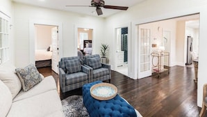 A spacious living room with a blue sofa, a plaid sofa, and a blue ottoman. There is a ceiling fan, a dining area with a table and chairs, and a kitchen area visible through an open door. - Our Nest at Spiller l Adorable Home near UA (Tuscaloosa)
