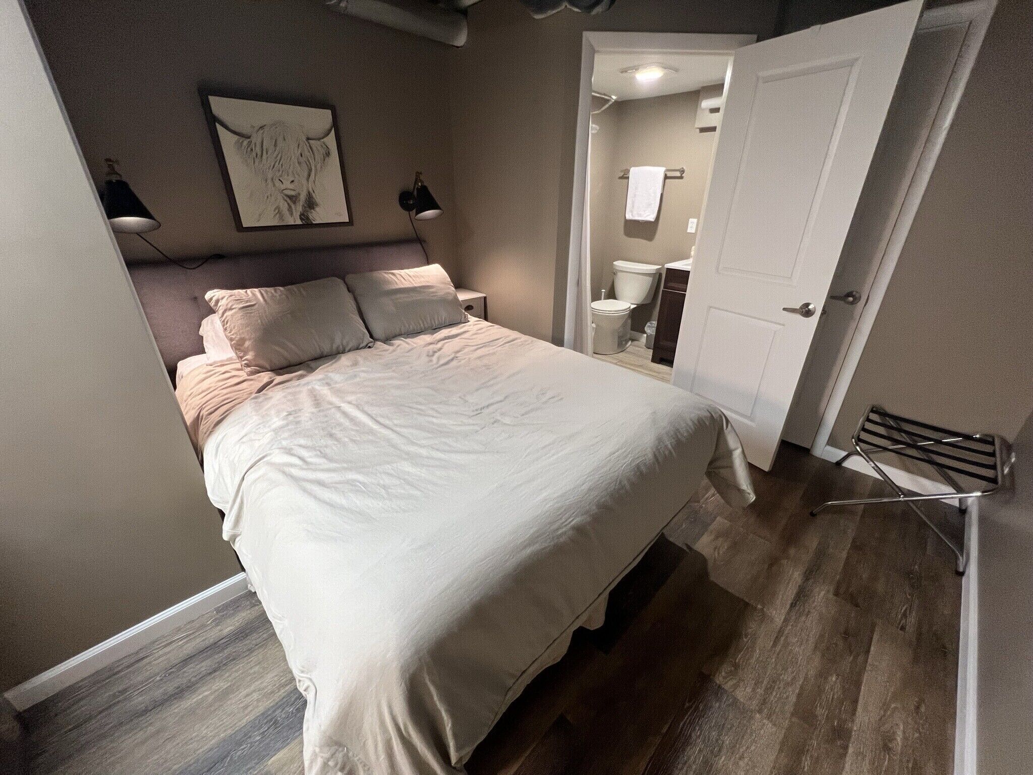 A bedroom with a bed, a nightstand, a lamp, a painting, and a bathroom visible through an open door.