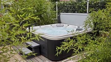 Un jacuzzi en un jardín con un deck de madera, rodeado de follaje y una valla metálica.