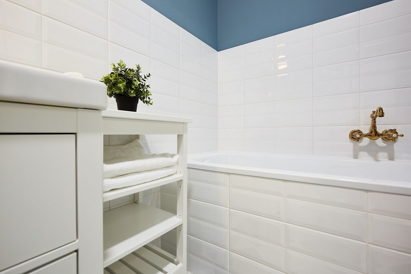 A bathroom with white tiled walls, a white bathtub with a gold faucet, and a shelf with a potted plant.