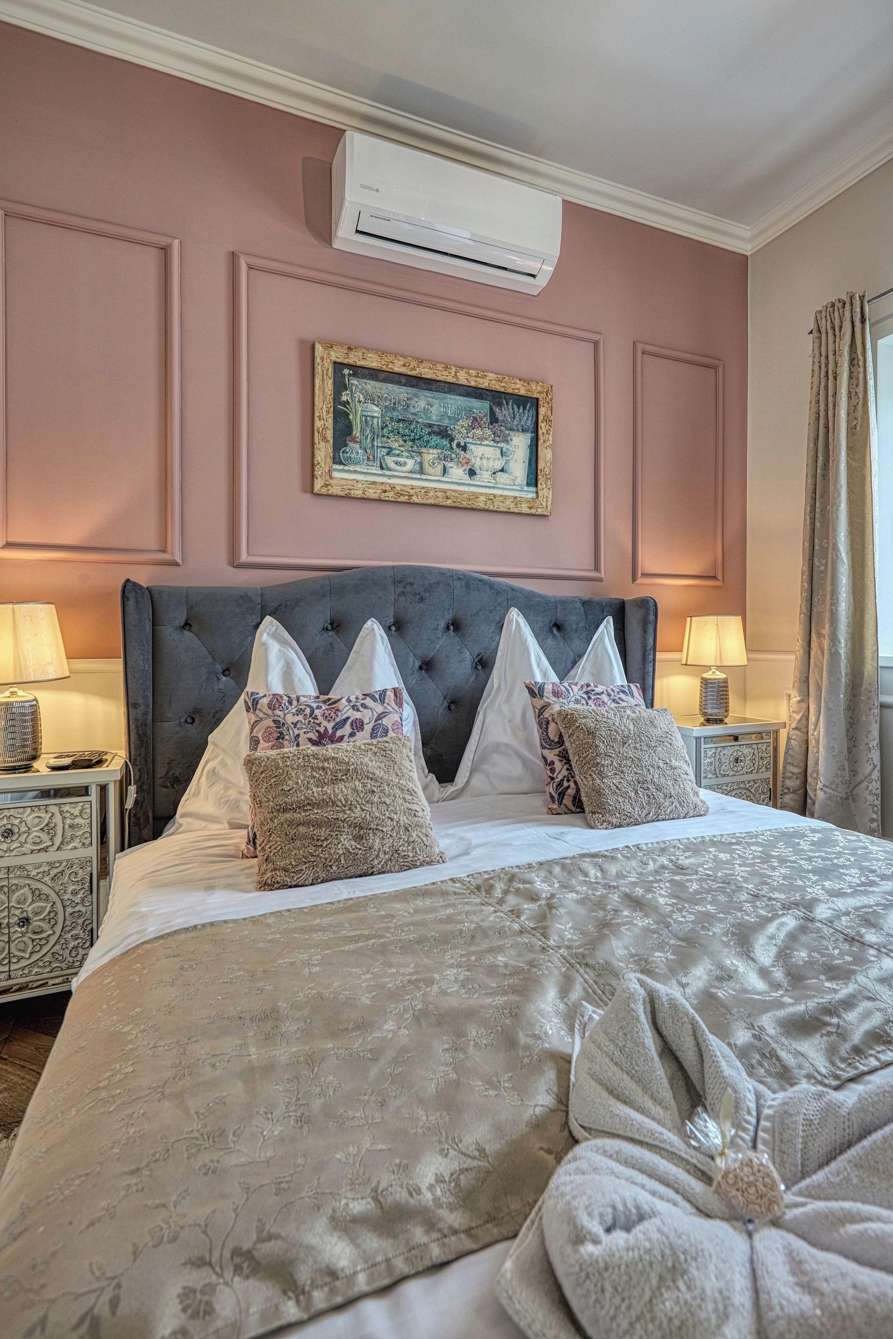 A neatly made bed with a decorative pillow, a bedside table with a lamp, and a wall-mounted air conditioner.