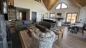 Een ruime woonkamer met een hoge plafond, een open haard, een bank, een gestreept fauteuil, en een eethoek met een tafel en stoelen.