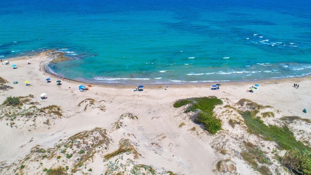 Vista aérea de una playa con aguas turquesas cristalinas, orilla arenosa y sombrillas dispersas.