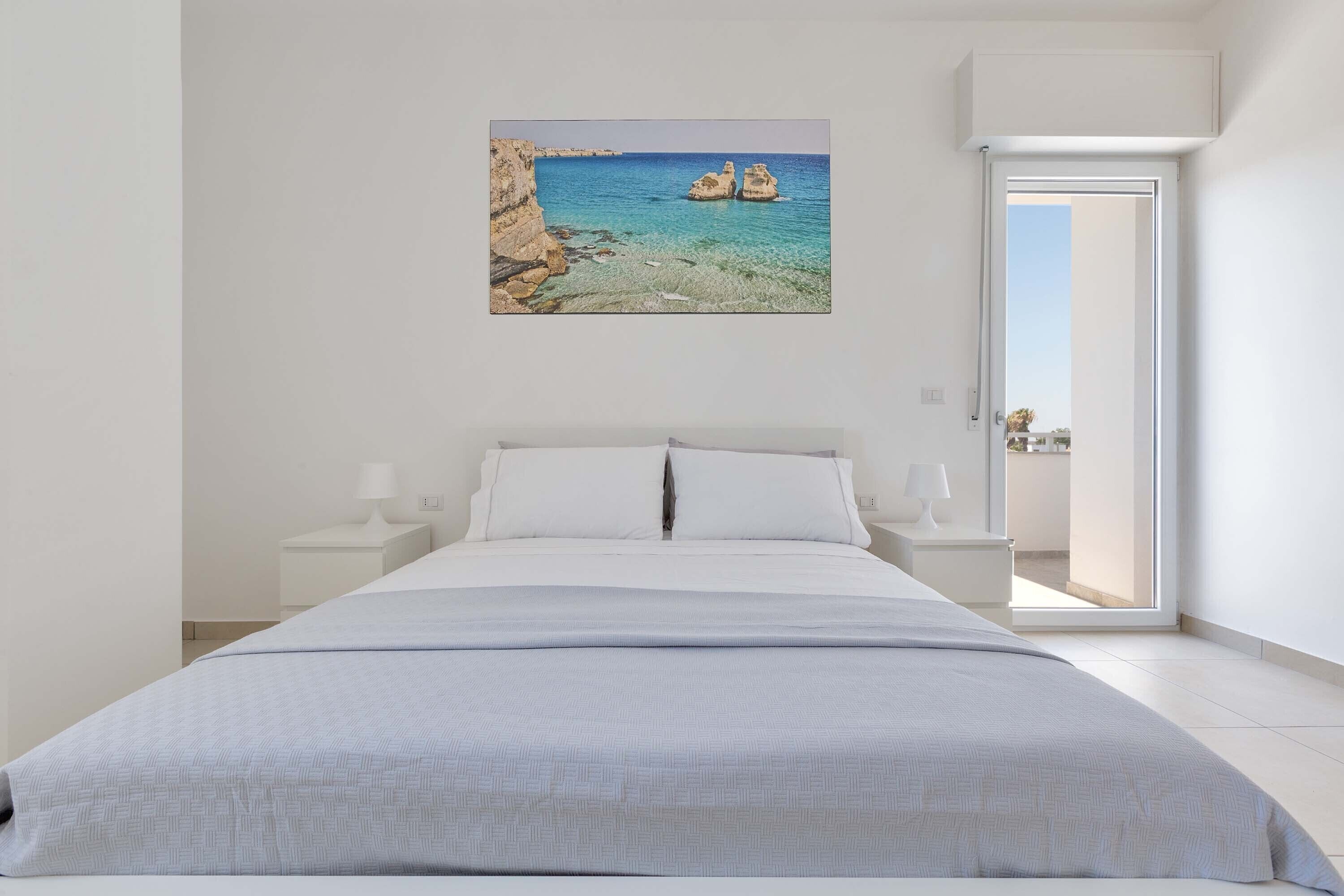 A bedroom with a bed, white nightstands, a painting of a coastal scene, and a door leading to an outdoor balcony.