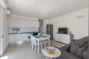 Una cocina moderna con armarios blancos, una mesa de comedor con sillas, un sofá gris y un televisor sobre un mueble.