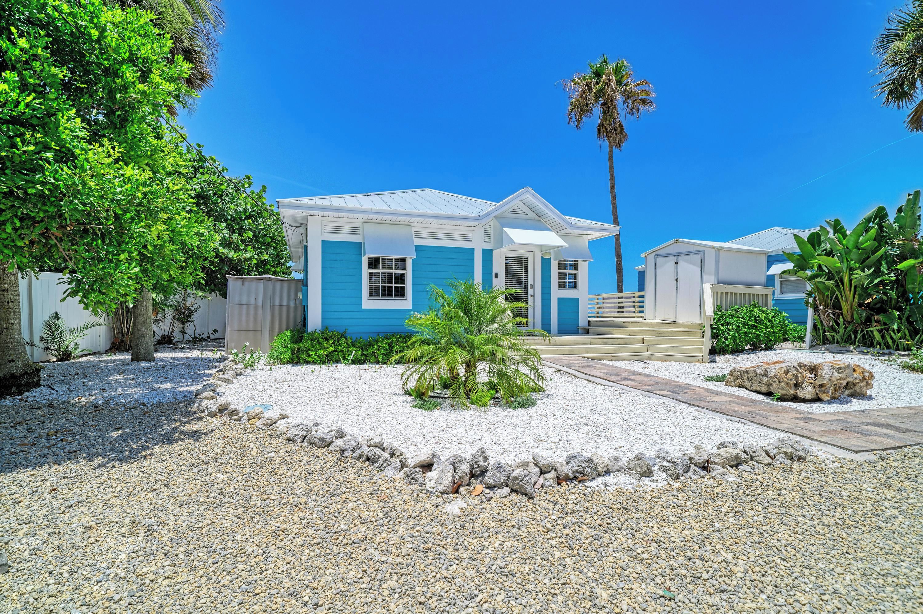 A two-story house with a blue exterior and a white roof, surrounded by a gravel driveway and tropical vegetation.